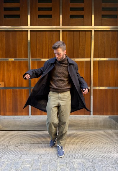 Hombre ajusta un abrigo negro abierto, lleva un suéter marrón de punto en cable, pantalones oliva y zapatillas azules, de pie contra una pared de paneles de madera.