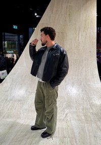 Fekete bőr rövid kabát, szürke pulóver, olíva színű zsebes katonai nadrág, és fekete cipő. Texturált háttér semleges árnyalatokkal.