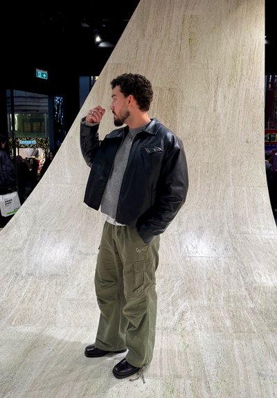 Chaqueta corta de cuero negro, suéter gris, pantalones cargo oliva con bolsillos y zapatos negros. Fondo texturizado con tonos neutros.