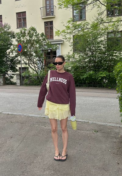 Sudadera marrón con el texto "WELLNESS", falda amarilla en capas, chanclas negras, sosteniendo una botella de agua amarilla, de pie en una acera.