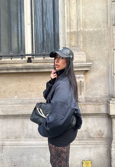 Joven con una chaqueta negra de gran tamaño, falda de encaje negra y gorra negra, sosteniendo un bolso negro, de pie junto a un edificio de piedra con una ventana con rejas.
