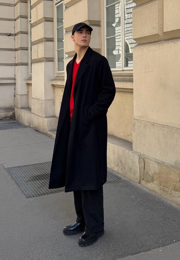 Cappotto lungo nero sopra una camicia rossa, abbinato a pantaloni neri e scarpe nere chunky. Il modello indossa anche un berretto nero. Sfondo urbano.