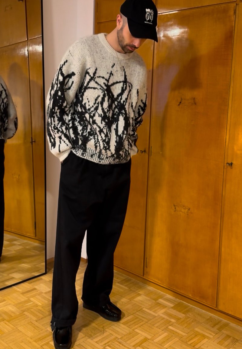 Pull en maille avec un motif abstrait noir sur fond crème, associé à un pantalon large noir et des chaussures noires. Casquette noire avec logo.