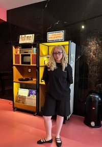 Melna īsām piedurknēm krekls un šorti uz personas ar teksturētām, sapītām matiem. Fona attēlā ir daudzkrāsains retro uzglabāšanas vienība ar vinila ierakstiem un televizoru.