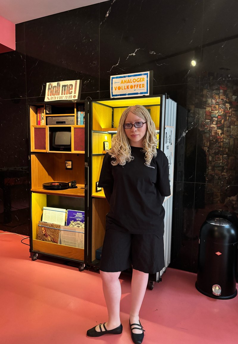 Black short-sleeve shirt and shorts on a person with textured, braided hair. A multicolored retro storage unit with vinyl records and a TV is in the background.
