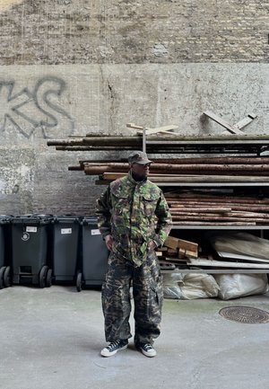 Hombre con chaqueta, pantalones y gorra de camuflaje de pie con las manos en los bolsillos junto a madera apilada y contenedores de basura negros contra una pared desgastada.