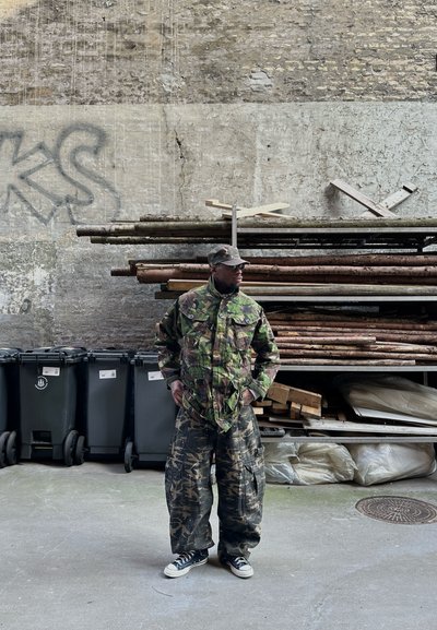Hombre con chaqueta, pantalones y gorra de camuflaje de pie con las manos en los bolsillos junto a madera apilada y contenedores de basura negros contra una pared desgastada.