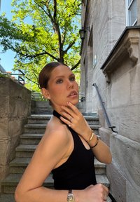 Vestido negro de cuello halter, de tela suave; modelo adornado con pulseras de oro, anillos y pendientes de perno; entorno natural con árboles verdes.