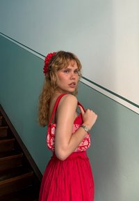 Czerwona sukienka z kwiatowymi detalami, dopasowana czerwona torebka z białym wzorem kwiatowym, falowane blond włosy, stojąca na schodach z niebieskozieloną ścianą.