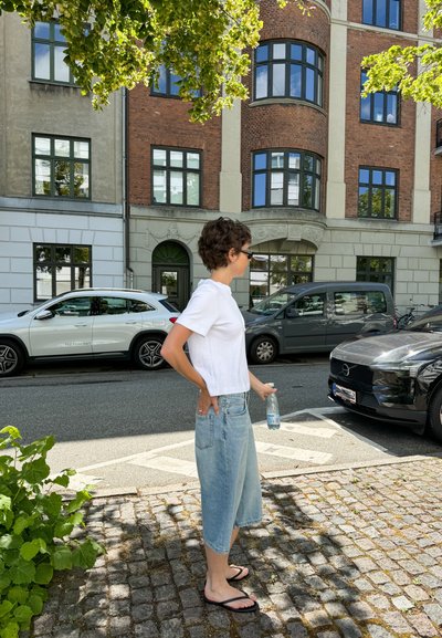 Camiseta blanca de manga corta, culottes de mezclilla azul claro, chanclas negras, sosteniendo una botella de agua, de pie junto a coches aparcados en una calle de adoquines.