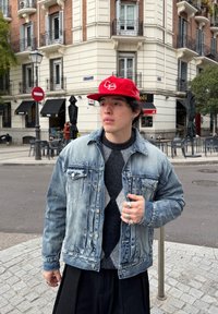 Jeune homme portant une casquette rouge, une veste en jean, un pull à motifs et des bagues, debout à un coin de rue urbain avec des cafés et des immeubles résidentiels en arrière-plan.