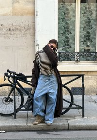 Jeans azuis oversized, top riscado em preto e branco, casaco longo castanho, sapatilhas de cano alto bege; de pé ao lado de uma bicicleta preta numa rua da cidade.