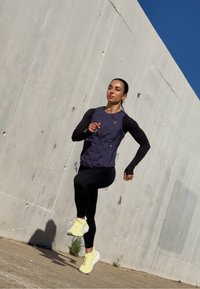 Lekkoatleta w granatowej kamizelce do biegania i czarnych legginsach, w żółtych butach do biegania, uchwycony w biegu na tle jasnej betonowej ściany.