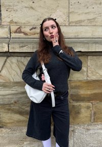 Pulôver de lã de gola alta em cinza escuro, combinado com calções de denim pretos. Uma mala de ombro branca e anéis de prata complementam o visual contra uma parede de pedra.