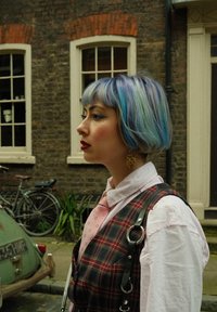 Καρό γιλέκο με άσπρο πουκάμισο, ροζ γραβάτα και χρυσά σκουλαρίκια. Μπλε και πράσινα μαλλιά, με ρινόδεσμο. Τοιχοποιία από τούβλα και ποδήλατα στο φόντο.