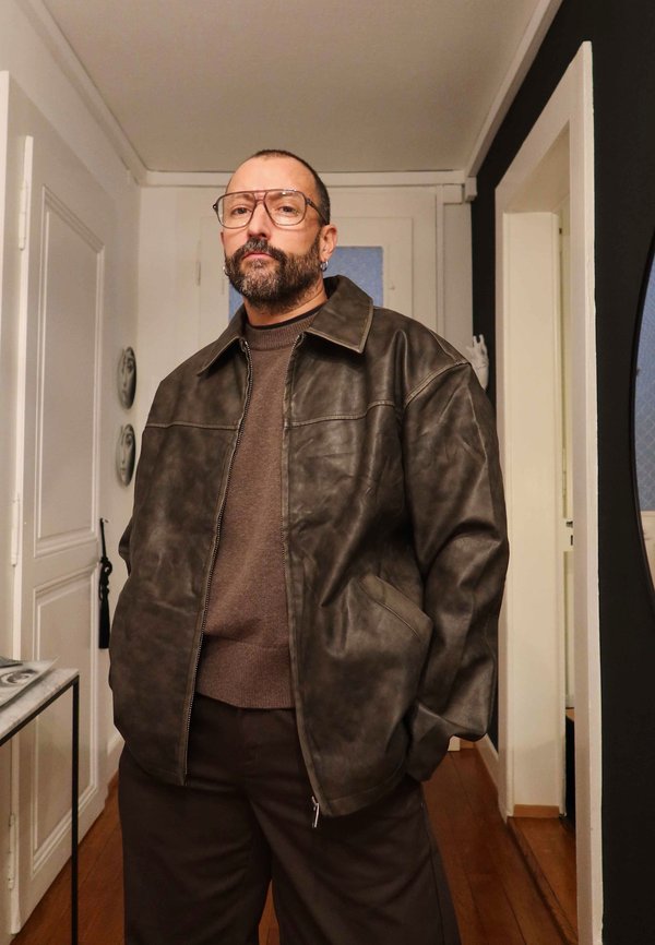 Brown leather jacket with a collared design, two front pockets, worn over a brown jumper and dark trousers, in a well-lit interior setting.