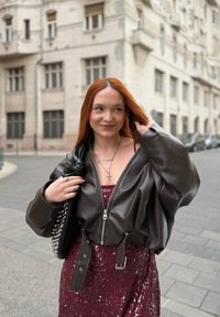 Braune Leder-Bomberjacke mit übergroßen Ärmeln, getragen über einem eng anliegenden burgunderfarbenen Pailletten-Kleid. Silberne Kettenhalskette und schwarze Handtasche mit Nieten.