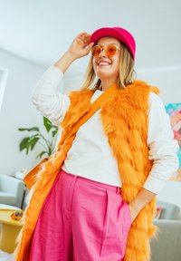 Χαμογελαστό άτομο που φοράει πορτοκαλί γιλέκο από ψεύτικη γούνα, λευκό μακρυμάνικο πουκάμισο, ροζ παντελόνι, ροζ καπέλο και πορτοκαλί ντεγκραντέ γυαλιά ηλίου μέσα σε εσωτερικό χώρο, με το χέρι στο καπέλο.