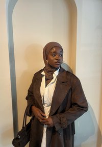 Palton trench din suede maro, purtat peste o cămașă albă, cu un hijab maro și cercei mari din aur, stând de-a lungul unui perete de culoare deschisă.