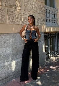 Denim korzet s čipkovým uzáverom vpredu, doplnený širokými čiernymi nohavicami. Štýl zahŕňa béžovú tašku a zlaté doplnky. Textúrované pozadie zo steny.