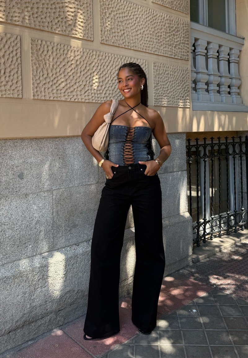 Top de corset em denim com frente com cordões, combinado com calças pretas de pernas largas. O estilo inclui uma bolsa bege e acessórios dourados. Fundo com parede texturizada.