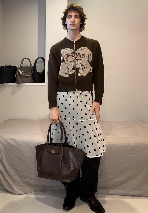 Junge Person trägt einen braunen Pullover mit Reissverschluss und Cartoon-Hunden, einen weißen Rock mit Punkten über schwarzen Hosen, hält eine große braune Handtasche.