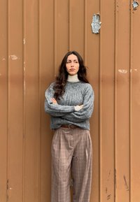 Femme aux longs cheveux bruns portant un pull gris et un pantalon large à carreaux, debout les bras croisés contre un mur en métal ondulé marron.