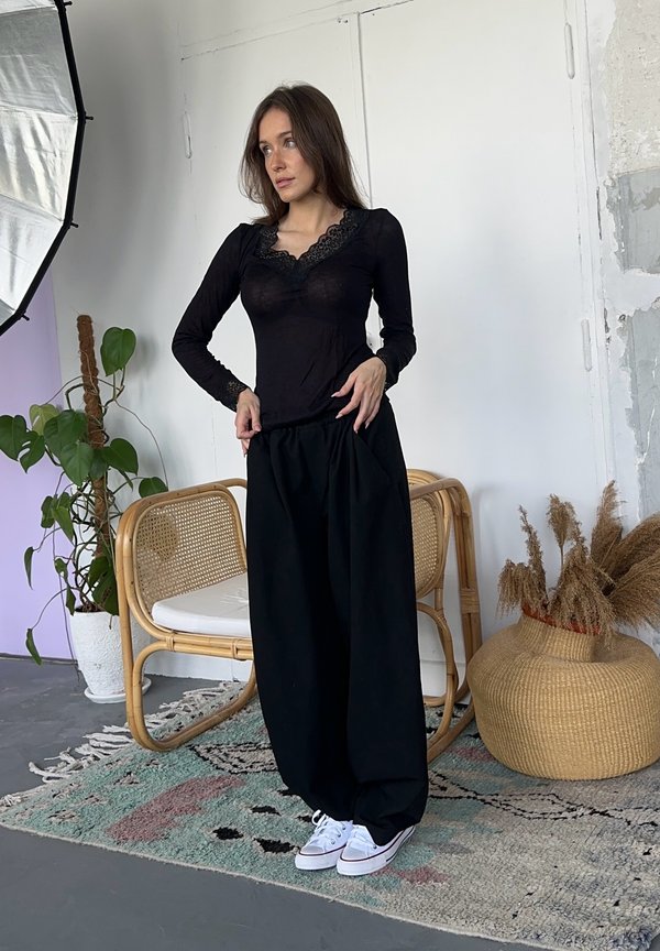 Haut noir à manches longues avec un col en dentelle, associé à un pantalon noir ample et des baskets blanches. Contexte intérieur avec des plantes et des accents en osier.