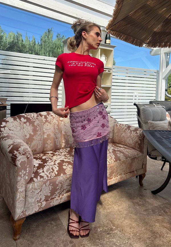 Jeune femme en chemise rouge avec texte et jupe violette posant à l'intérieur près d'un canapé à fleurs et de mobilier de patio extérieur.