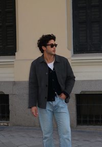 Casaco cinza em mistura de lã sobre um cardigan preto, combinado com calças de ganga azul claro. Acessórios incluem óculos de sol pretos. Cenário ao ar livre com parede.
