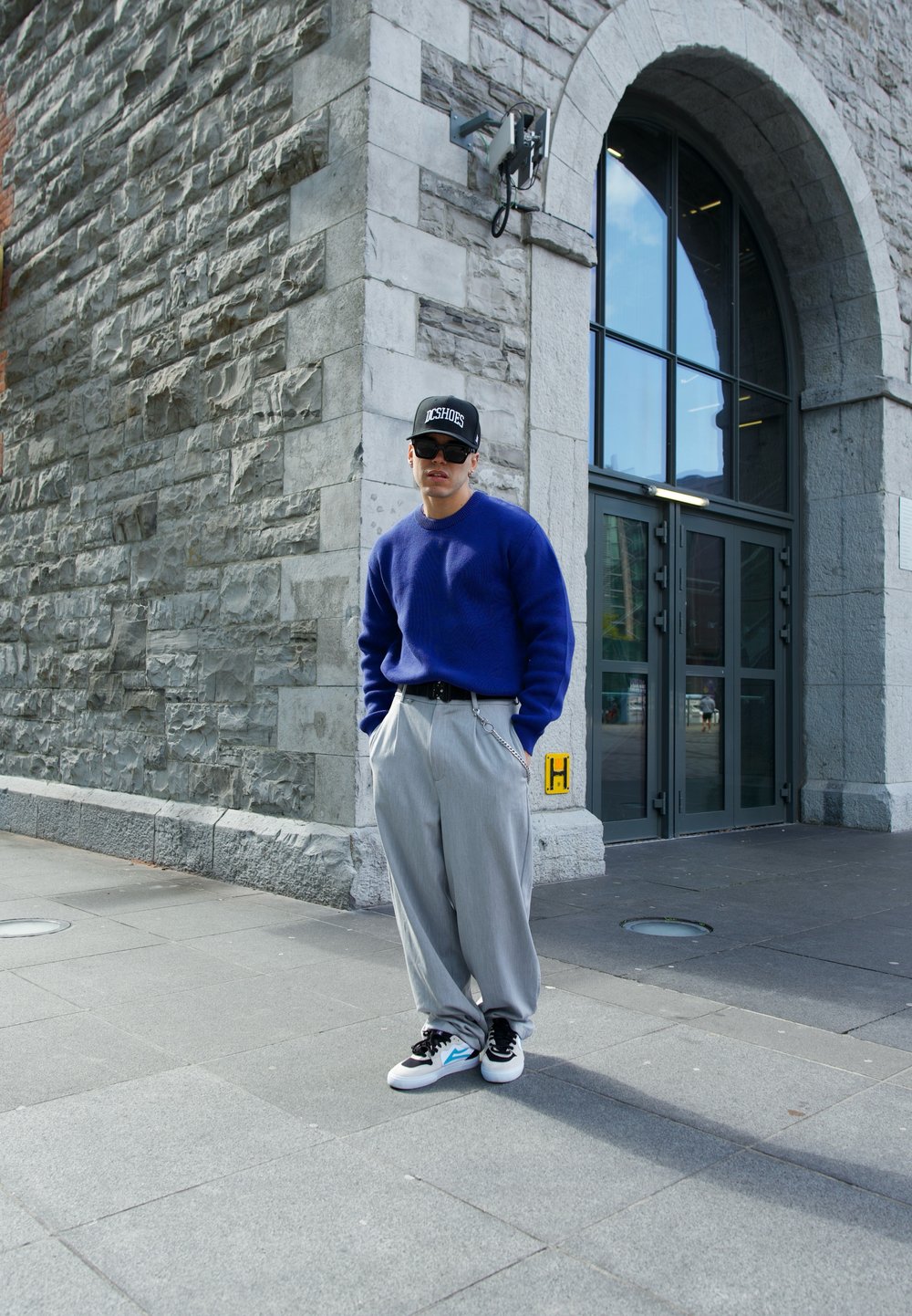 Blå strikket sweater, grå vidde bukser, sorte sneakers med teal accenter. Tilbehøret inkluderer en sort kasket med hvid tekst. Urban baggrund.