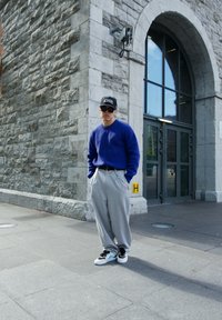 Blå strikket sweater, grå vidde bukser, sorte sneakers med teal accenter. Tilbehøret inkluderer en sort kasket med hvid tekst. Urban baggrund.