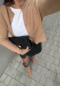 Cardigan tricotat maro, cămașă albă, poșetă neagră cu accesorii aurii, fustă neagră și cizme bej până la genunchi pe o suprafață pavata.