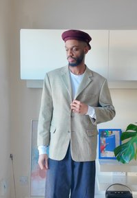 Beige checked blazer with notched lapels, two front pockets, layered over a white shirt, paired with dark trousers, and a maroon hat.