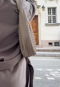 Šviesiai ruda audinė odinė rankinė su tekstūriniu paviršiumi, dėvima kartu su pilku paltu, dalinai matoma su medinėmis durimis fone.