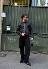 Μαύρο δερμάτινο μπουφάν με κλείσιμο φερμουάρ, συνδυασμένο με μαύρο φαρδύ παντελόνι και μαύρα παπούτσια. Αξεσουάρ περιλαμβάνουν γυαλιά ηλίου και μια μικρή τσάντα.