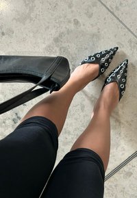 Sorte spidse sko med nitteudstyr, kombineret med en sort læderhåndtaske. Der bæres leggings, og gulvet er lyst.