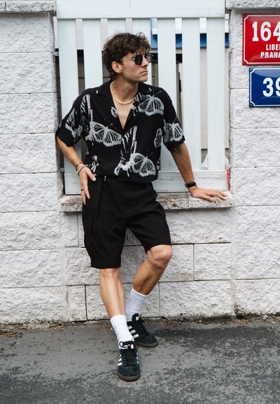 Camisa negra de manga corta con estampado de mariposas blancas, pantalones cortos negros, calcetines blancos y zapatillas negras. Los accesorios incluyen gafas de sol y un collar de perlas.