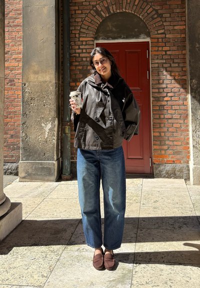 Joven mujer con gafas, que lleva una chaqueta oscura y vaqueros azules, sostiene una taza de café con estampado, de pie frente a una puerta roja y una pared de ladrillos.