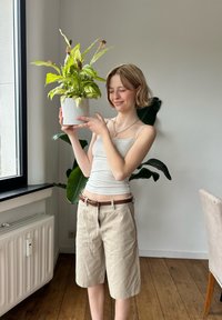 Potted green plant with broad leaves in a textured white pot. A person is holding the pot, dressed in a light tank top and beige shorts.