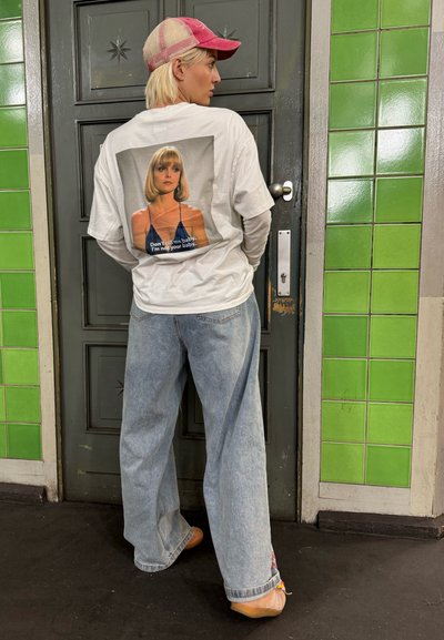 Camiseta blanca de manga larga con un estampado gráfico en la espalda, combinada con jeans de pierna ancha de color azul claro y una gorra rosa. Fondo de azulejos verdes.