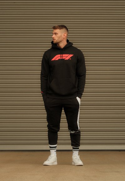 Sudadera negra con logo rojo, pantalones de chándal negros a juego con rayas blancas y zapatillas blancas. Textura suave, corte casual, diseño deportivo.