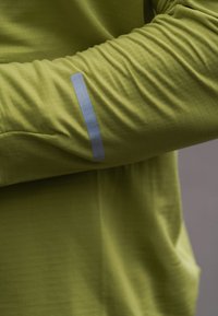 Långärmad olivgrön skjorta med en vertikal grå reflexstrimma på armbågsområdet mot en enkel bakgrund.