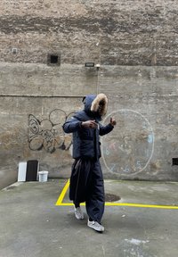 Námořnická modrá péřová bunda s kapucí podšitou kožešinou, zkombinovaná s volnými černými kalhotami a kostkovanými slip-on botami, na pozadí texturované zdi.