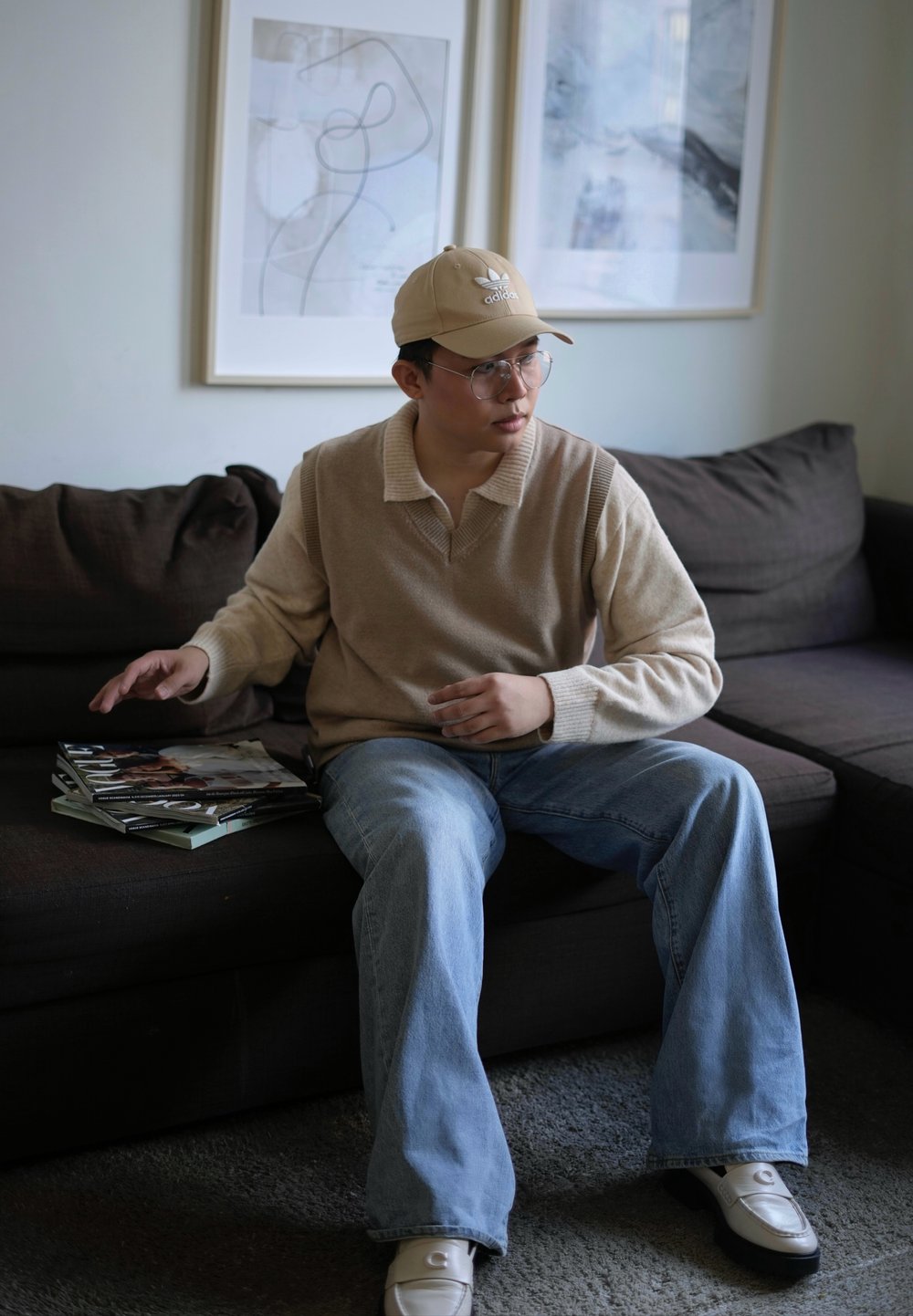 Beige collared sweater, light blue jeans, cream loafers with an accent, clear glasses, and a beige cap. A stack of magazines on a dark sofa.