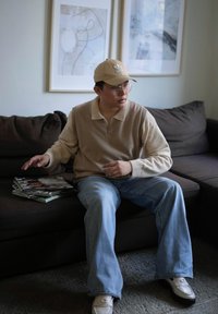 Beige collared sweater, light blue jeans, cream loafers with an accent, clear glasses, and a beige cap. A stack of magazines on a dark sofa.
