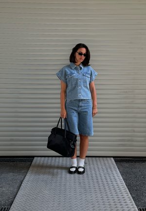Conjunto de dos piezas de denim que incluye una blusa de manga corta con botones y pantalones cortos hasta la rodilla. Bolso oscuro, calcetines blancos y zapatos negros.