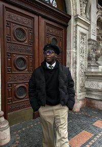 Sort puffer jakke, sort crewneck sweater, beige bukser og en sort béret. Står foran udsmykkede trædøre med indviklede udskæringer.