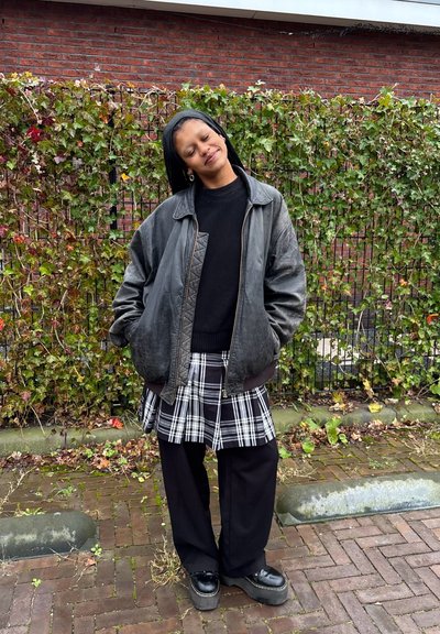 Chaqueta bomber de cuero negro con detalle acolchado, suéter negro, falda de cuadros y pantalones anchos negros. Combinada con zapatos de plataforma gruesos.