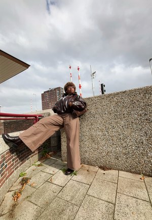Chaqueta marrón brillante, pantalones de pierna ancha beige, botas oscuras; posada contra una pared de concreto texturizada con postes a rayas rojas y blancas de fondo.